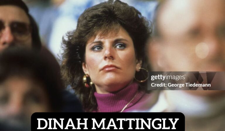 Dinah mattingly with long blonde hair smiles warmly, standing outdoors near trees. She is dressed in a black top and wears a silver necklace.