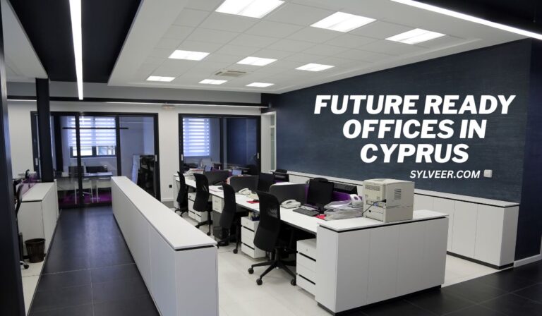 Modern office interior with sleek white desks, black chairs, and a printer, featuring a large sign on a dark wall that reads “FUTURE READY OFFICES IN CYPRUS – SYLVEER.COM.” The space has glass-walled private offices and bright overhead lighting.